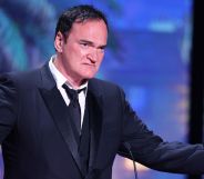 Quentin Tarantino on stage speaking in a black suit and tie, holding his arms wide.