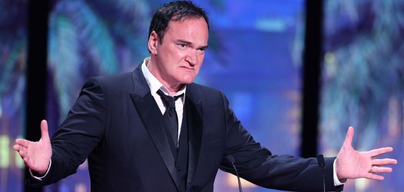 Quentin Tarantino on stage speaking in a black suit and tie, holding his arms wide.