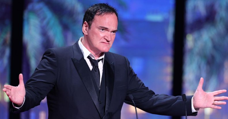 Quentin Tarantino on stage speaking in a black suit and tie, holding his arms wide.