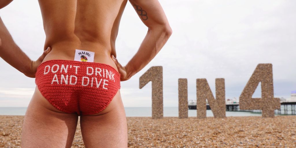 Tom Daley in Brighton Beach posing in red knitted swim trunks that read Don't Drink and Dive