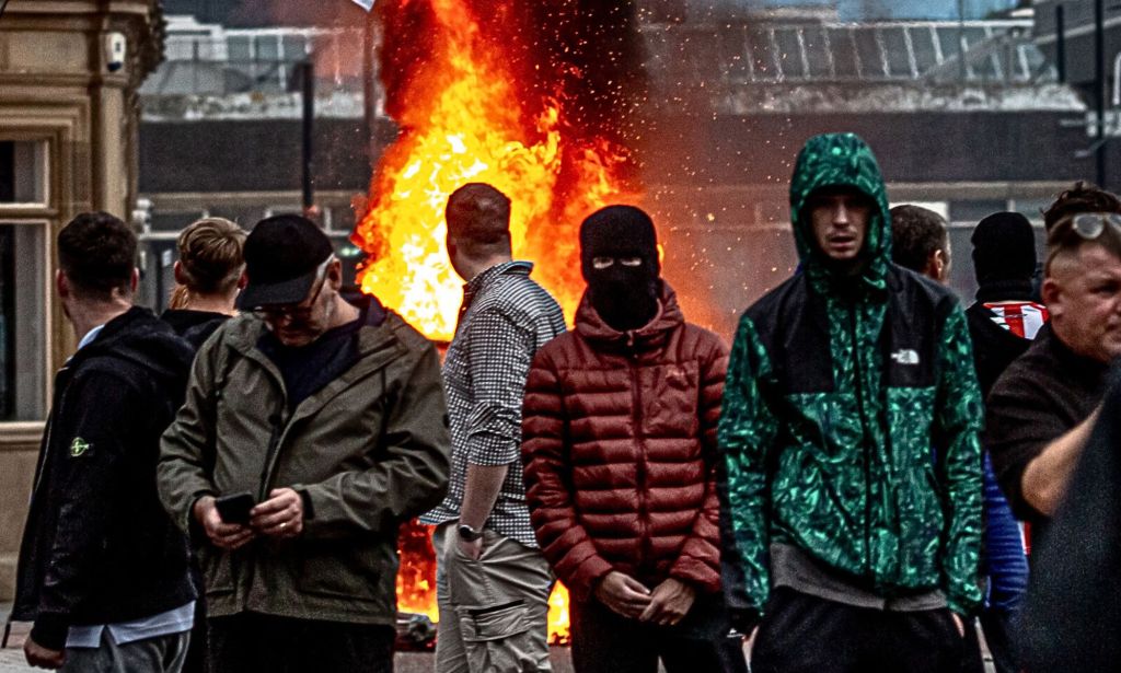 A group of far-right thugs stand surrounding a fire.