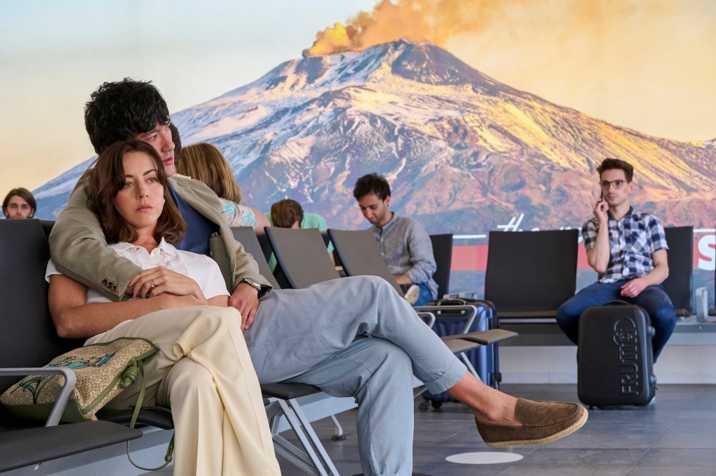 Aubrey Plaza and Will Sharpe leaning on each other in an airport in The White Lotus with an erupting volcano behind them on a screen