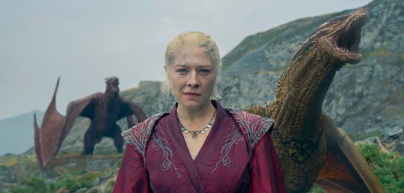 Rhaenyra dressed in a red robe with 2 roaring dragons behind her.