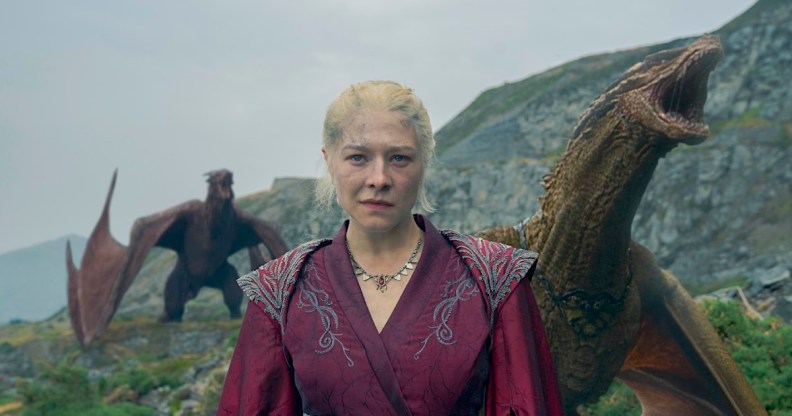 Rhaenyra dressed in a red robe with 2 roaring dragons behind her.