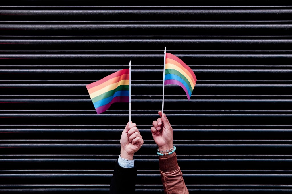 The hands of two Latino men come together to hold a vibrant multicolored flag, symbolizing their celebration and support of LGBTQ+ pride, set against the backdrop of a closed shop door.