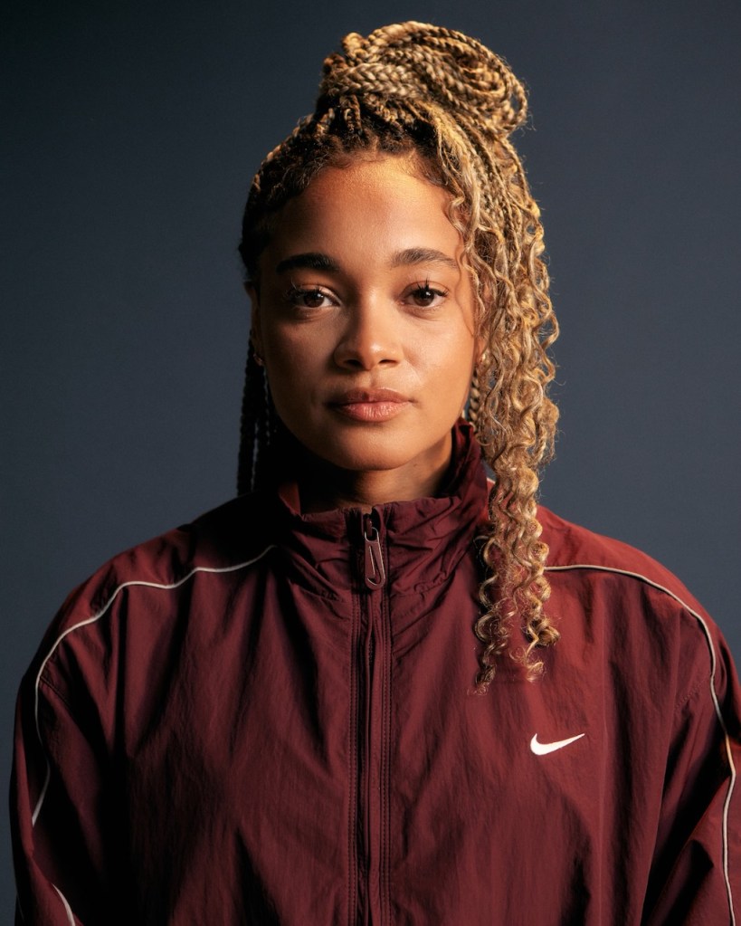 This is an image of a Black woman, British Olympian Alice Dearing. She is wearing a maroon Nike tracksuit. She has long brown hair. She is looking directly into the camera. 