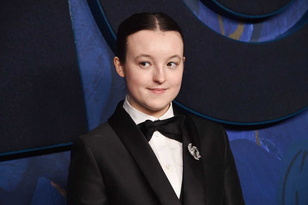 Bella Ramsey in a white shirt and black suit and bow tie. Their hair is tied back and they are looking to the right.