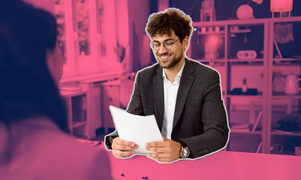 This is an image of a man wearing a charcoal blazer and a white shirt. He has curly short brown hair, a beard and is wearing glasses. He is smiling and is holding a piece of paper. The background is a blurred out meeting room and is stylised in fuschia.