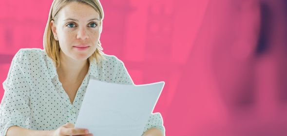 This is an image of a white woman with shoulder length blond hair holding a piece of paper. The background is a blurred out meeting room and is stylised in fuschia.