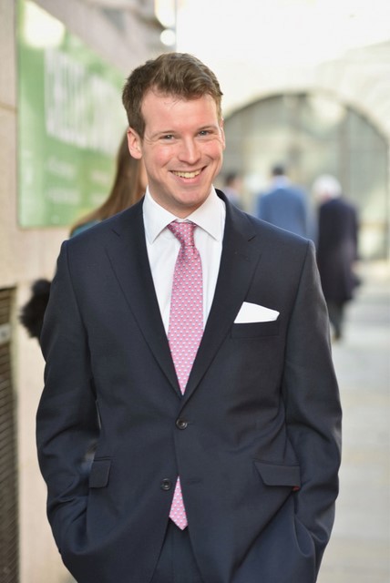 This is an image of Chris Hadrill. He is wearing a dark suit, white shirt and pink tie. He is smiling and his hands are in his pockets