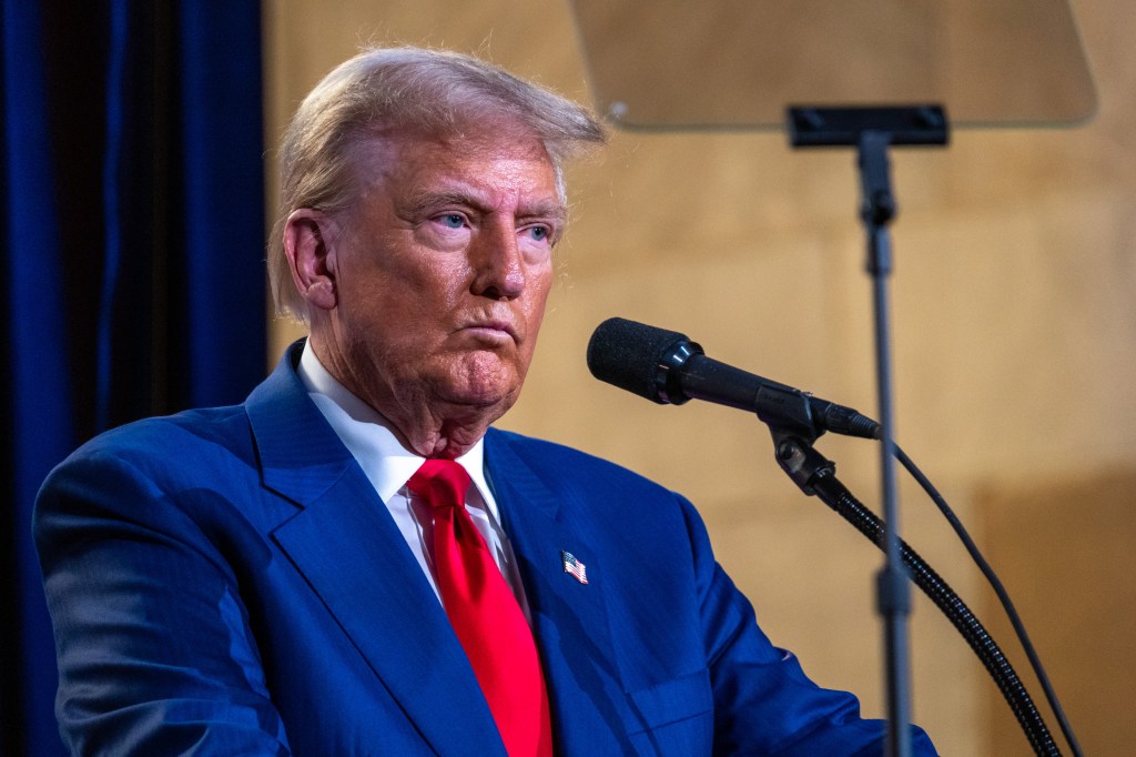 Donald Trump in a navy suit, white shirt and red tie standing at the mic looking annoyed.