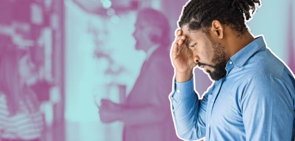 This is an image of a Black man looking frustrated while leaning on a wall. He has short dread locks tied up behind his head. He has a beard and is wearing a blue shirt.