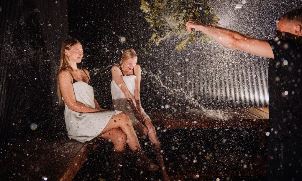 A sauna master shaking leaves at two individuals sitting in an Estonian sauna.