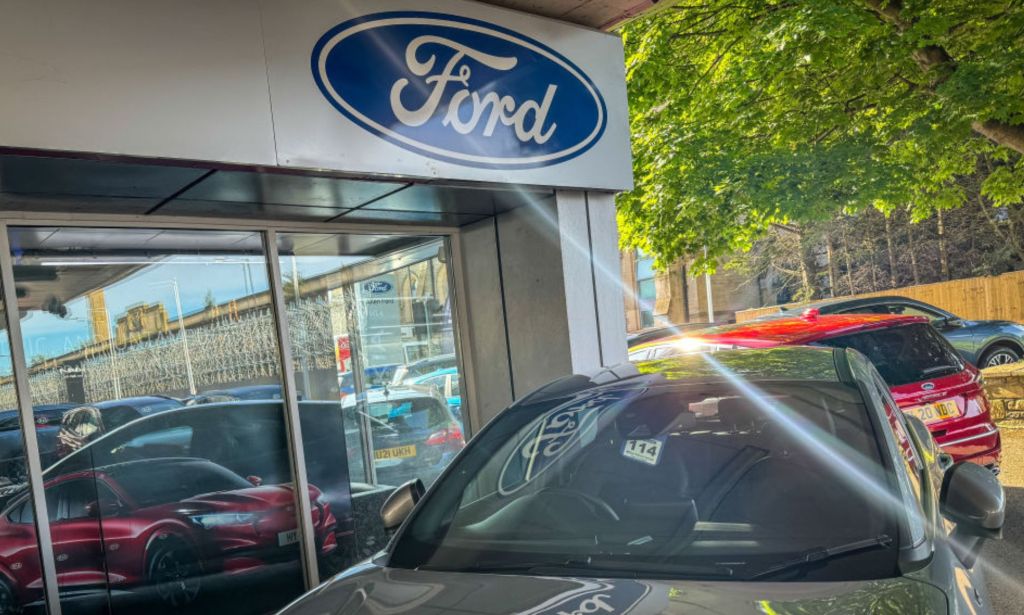 This is an image of a Ford car dealership. The iconic blue with silver lettered FORD logo is on a glass building. There are a few cars in the photo as well.