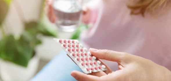 Femme-presenting person holding a pack of contraceptive pills and a glass of water.