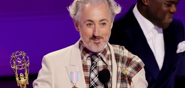 Alan Cumming on stage at the Emmys, holding his award, close cropped to show the trans pride flag medal