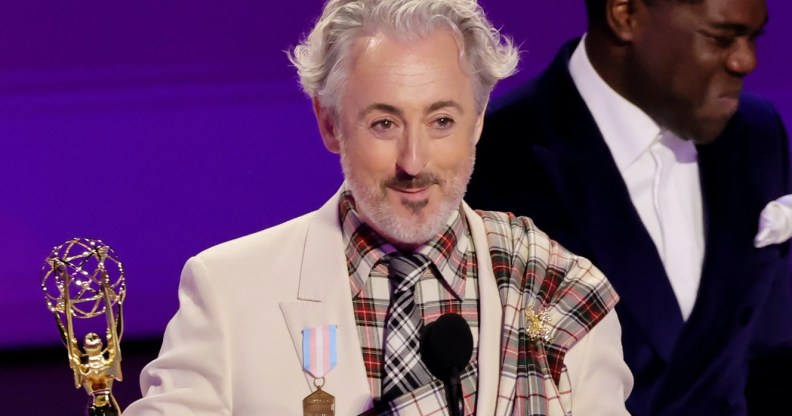 Alan Cumming on stage at the Emmys, holding his award, close cropped to show the trans pride flag medal