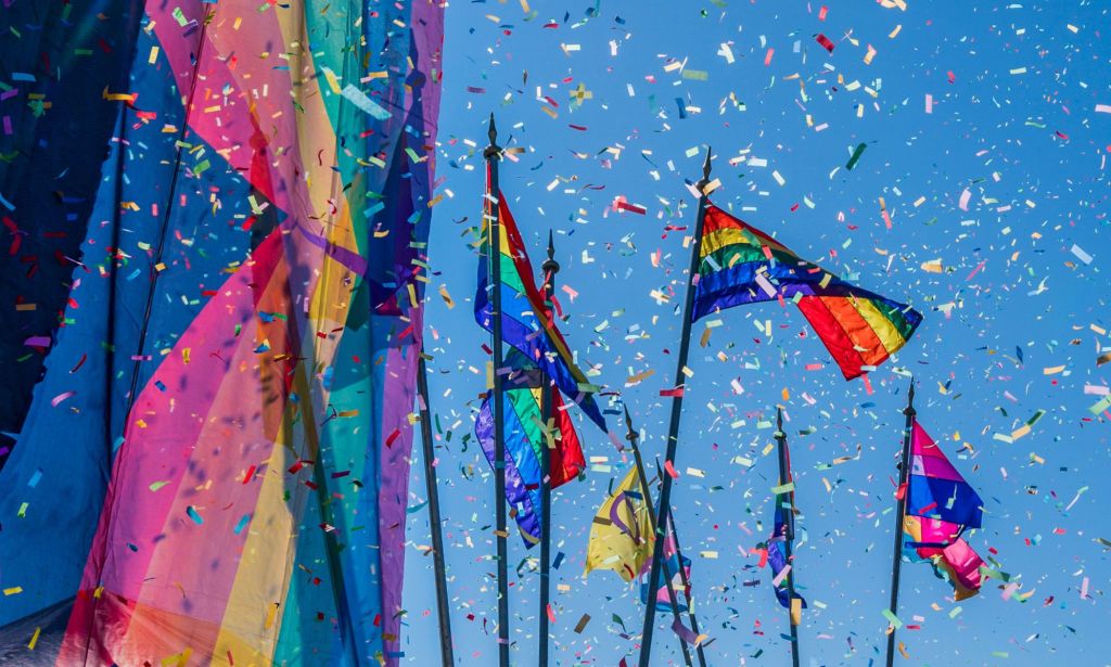 This is an image of confettie flying amongst large Pride flags on a sunny day. 