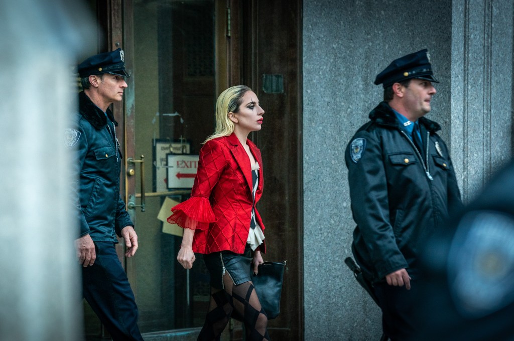 Harley Quinn exiting a building between two police men, she is wearing a red blader and black and white Joker make-up