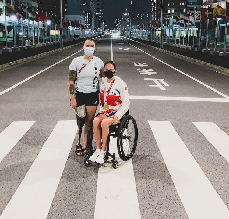 This is an image of two women standing in a crosswalk. One is in a wheelchair and the other woman wears a prosthetic leg.