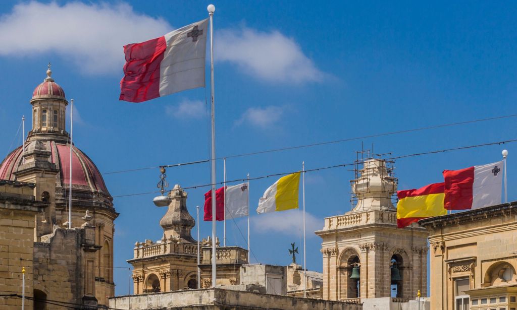 This is an image of a town in Malta. There are 3 Maltese flags in red and white and several sandstone type buildings.