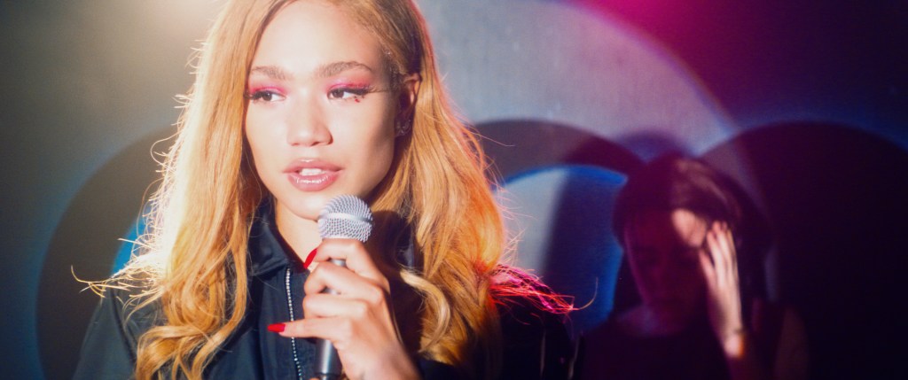 Queens of Dram still, a young woman is standing on stage in the spotlight with a mic in hand