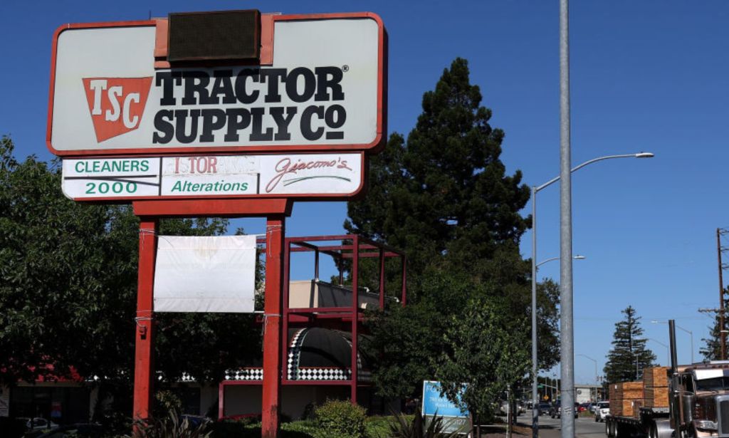 This is an image of a sign for the store Tractor Supply Co. It is in a rural setting and there are large pine trees in the background.