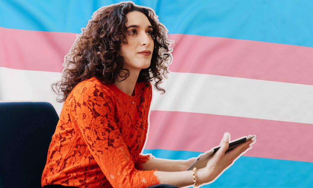 This is a transgender woman sitting on chair. Behind her is a graphic of the trans pride flag. 