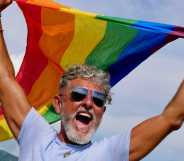 Portrait of a gray-haired elderly Caucasian man with a beard and sunglasses holding a rainbow LGBTQIA flag against a sky background, shouts in protest, Celebrates Pride Month Coming Out Day