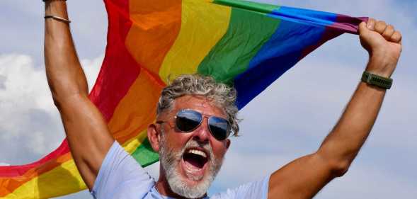 Portrait of a gray-haired elderly Caucasian man with a beard and sunglasses holding a rainbow LGBTQIA flag against a sky background, shouts in protest, Celebrates Pride Month Coming Out Day