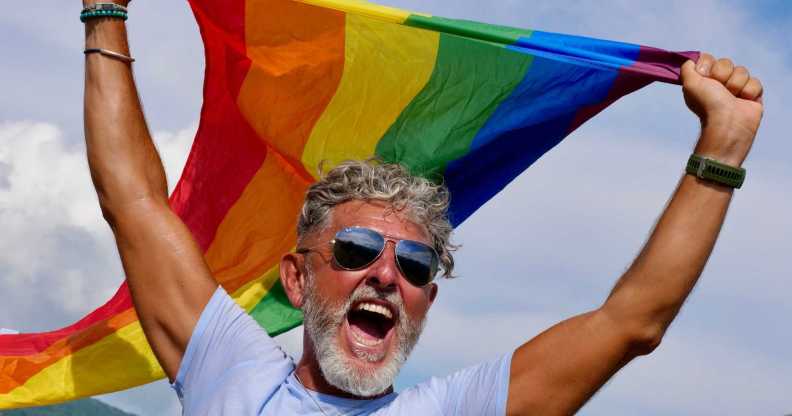 Portrait of a gray-haired elderly Caucasian man with a beard and sunglasses holding a rainbow LGBTQIA flag against a sky background, shouts in protest, Celebrates Pride Month Coming Out Day