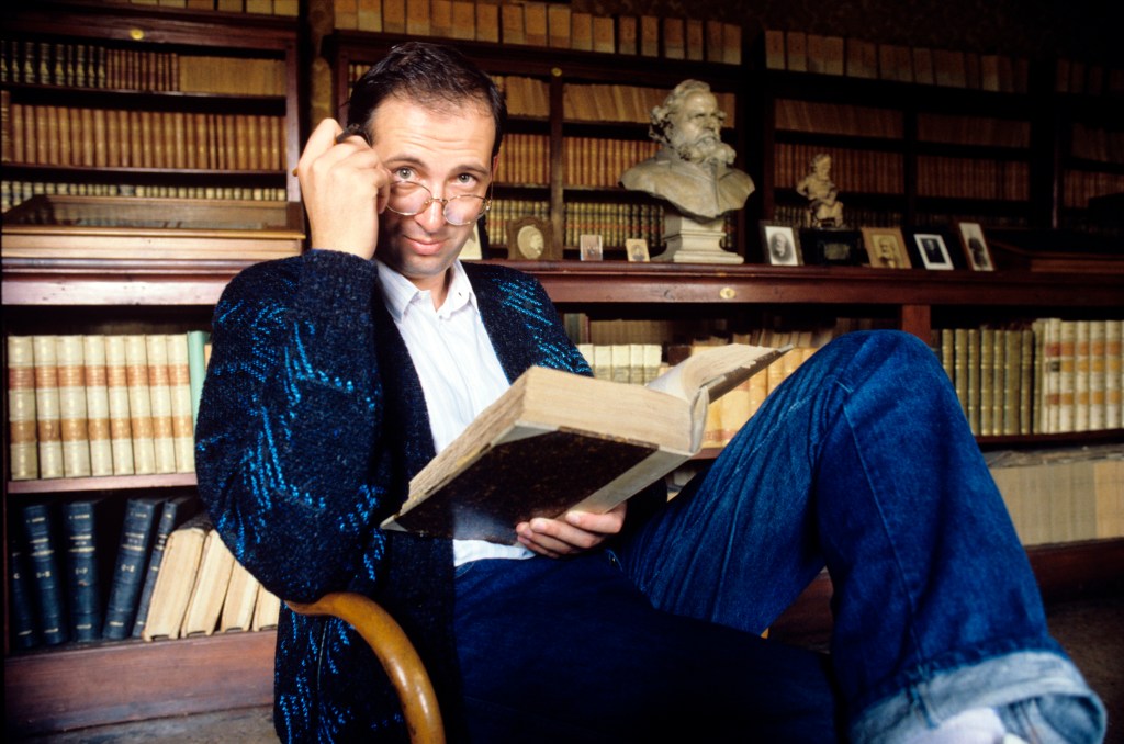 Italian writer Pier Vittorio Tondelli adjusts his glasses while reading a book, sitting on an armchair; behind him a shelf of his furnished library