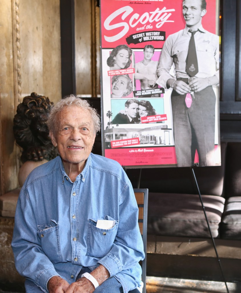 Scotty Bowers smiling in a denim blue shirt sat in front of the Scotty Bowers documentary poster