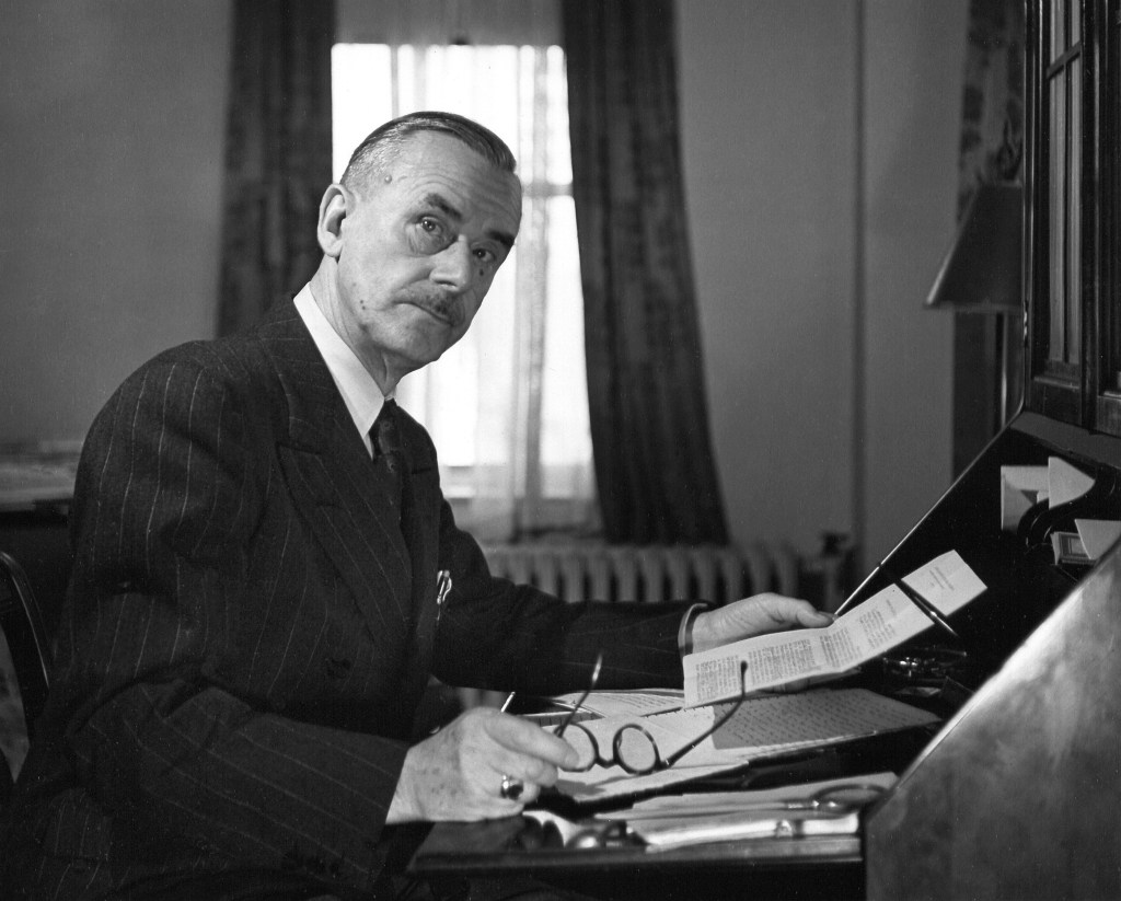 Portrait of German author Thomas Mann as he sits at his desk, a piece of paper in one hand and his glasses in the other