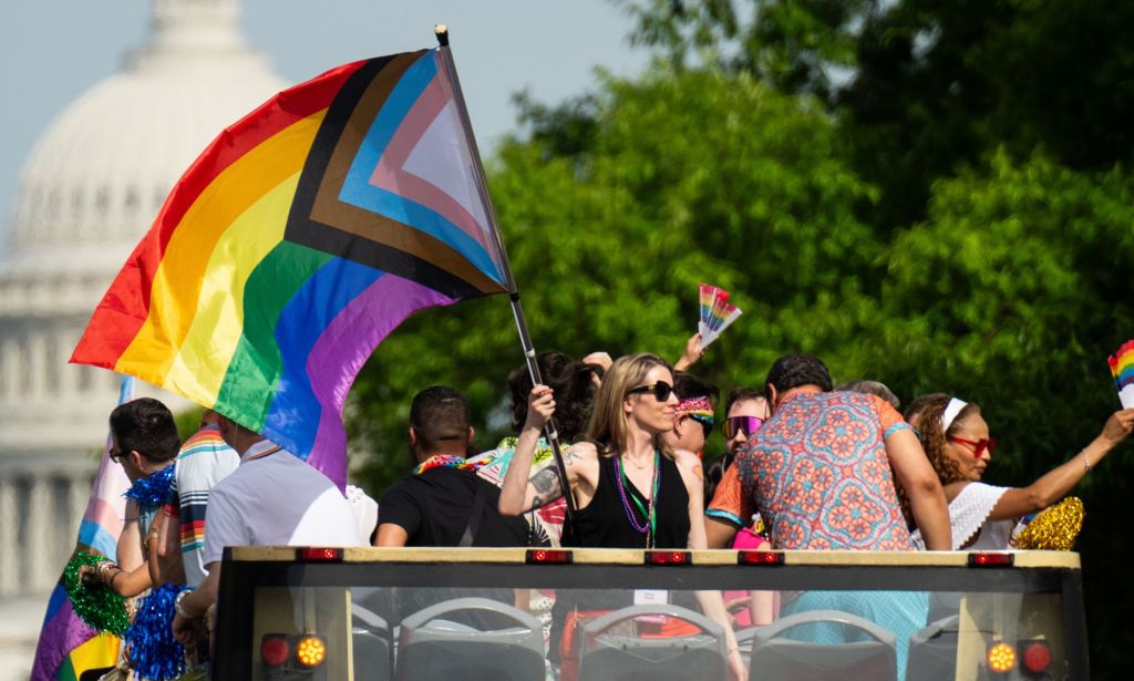 Someone waving an LGBTQ+ flag infront of the US Capitol building.