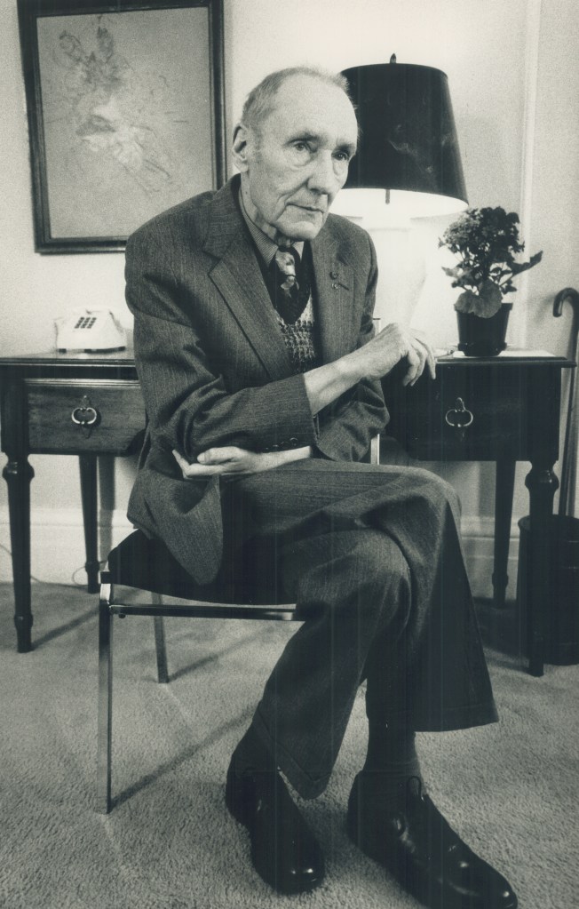 American author William Burroughs, caught in a reflective moment, sitting at a desk smoking