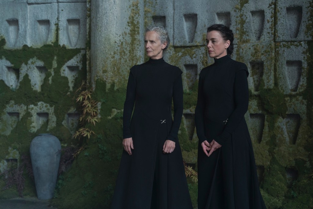 Barbra Marten and Olivia Williams in Dune: Prophecy. Dressed in full black against a mossy wall.