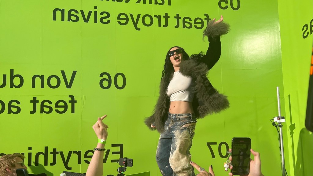 Charli XCX shared her new remix album with a few hundred fans at a special event. Huge lime green wall behind charli in distressed jeans, cropped white top and fuzzy jacket.