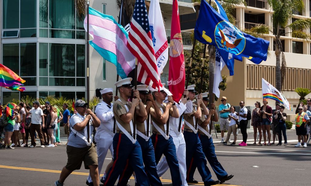 US military service members holding a variety of flags, including the US flag and the trans flag.