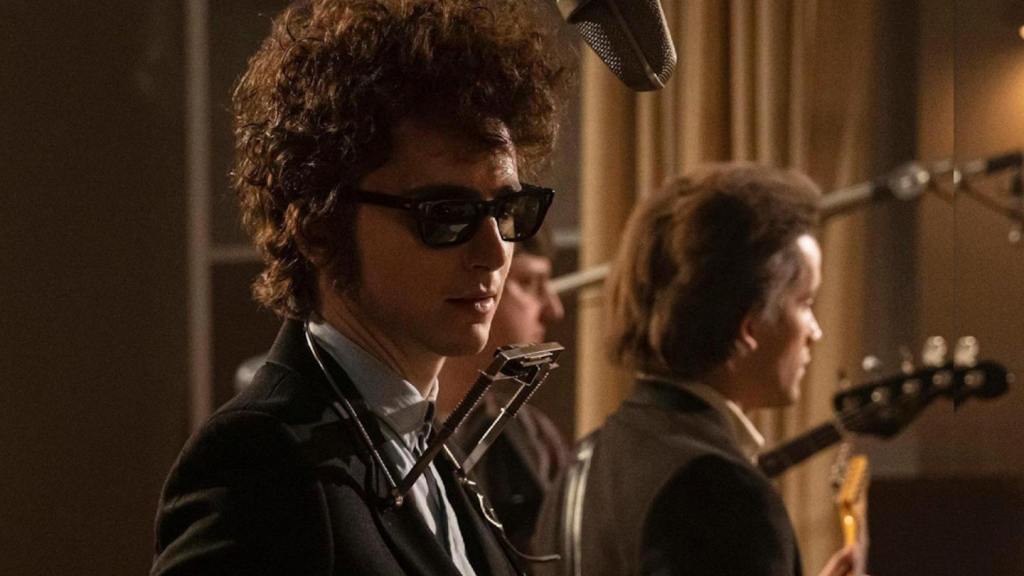 A Complete Unknown still: Timothée Chalamet as Bob Dylan in a studio recording. He's got big curly hair, dark sunglasses and a harmonica stand around his neck.