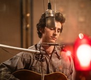 A Complete Unknown still: Timothée Chalamet as Bob Dylan in a studio recording. He's got a harmonica stand around his neck and playing the guitar.