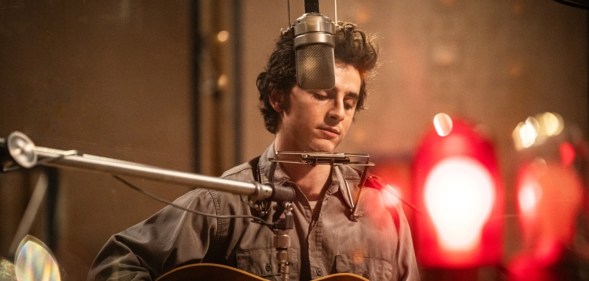 A Complete Unknown still: Timothée Chalamet as Bob Dylan in a studio recording. He's got a harmonica stand around his neck and playing the guitar.