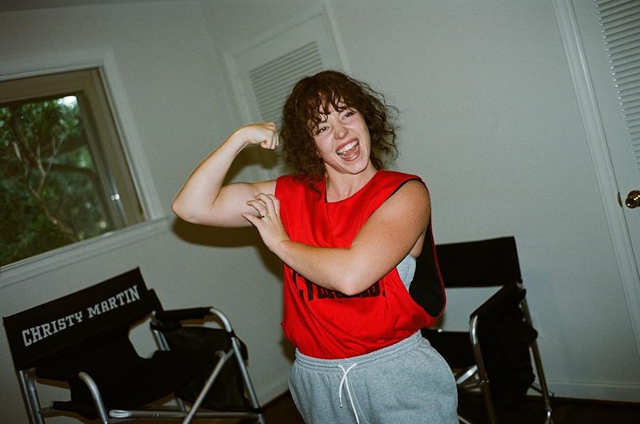 Sydney Sweeney on set of the Untitled Christy Martin Biopic; wearing grey sweatpants and a red vest, flexing her bicep.