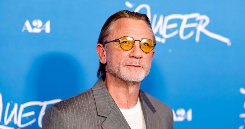 Daniel Craig in front of a blue background wearing orange lens glasses, a grey suit and white t-shirt