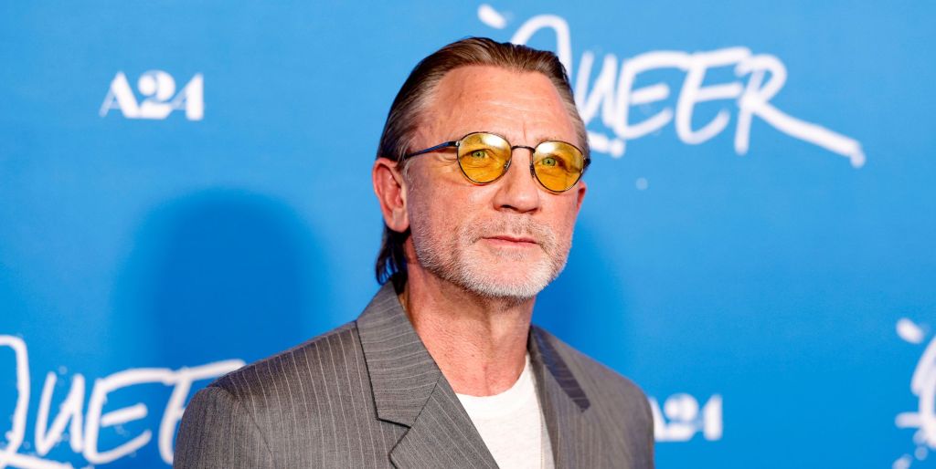 Daniel Craig in front of a blue background wearing orange lens glasses, a grey suit and white t-shirt