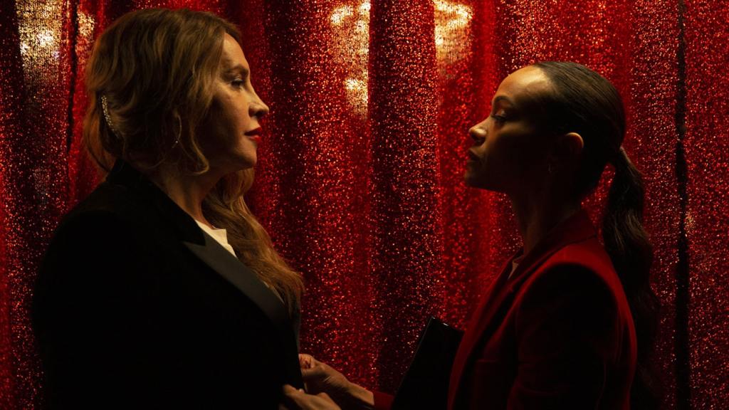 Emilia Pérez still: Karla Sofía Gascón and Zoe Saldana stood face to face behind a red glittering curtain.