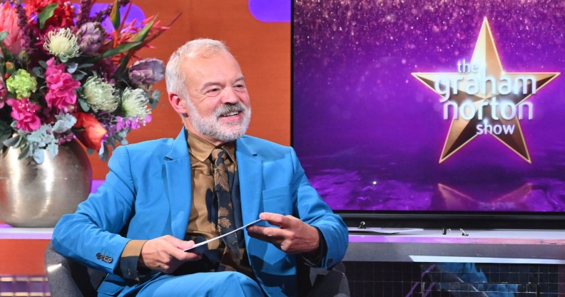 The Graham Norton Show, Graham Norton, in a blue suit, sitting next to a screen with the show name.