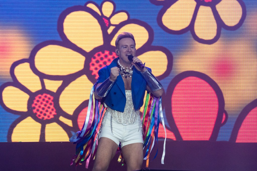 Ian "H" Watkins of Steps performs during the Brighton and Hove 2023 Pride. H is performing with big cartoon flowers behind him. He's wearing a silver-white corset and shorts with a blue waistcoat decorated with Pride flag coloured streamers.