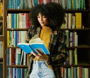 A person in front of an array of books reading from the pages of a paperback.