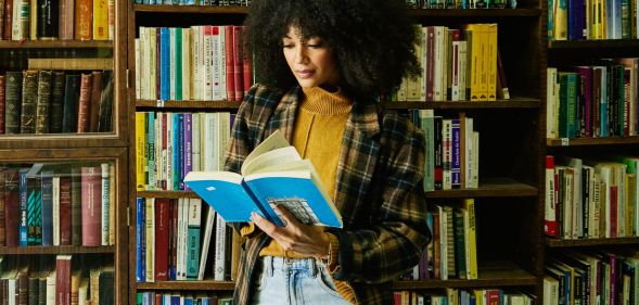 A person in front of an array of books reading from the pages of a paperback.